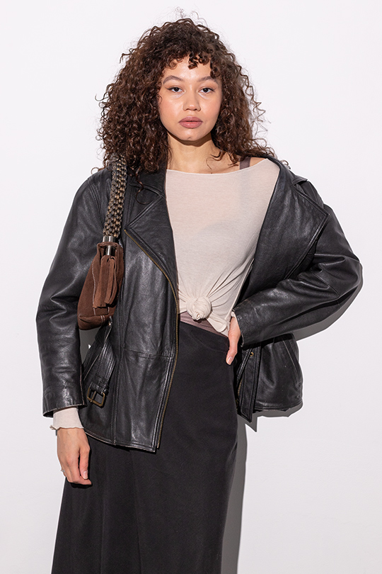 На фото кудрявая девушка в черной куртке и бежевом полупрозрачном лонгсливе с коричневой замшевой сумкой на плече. 